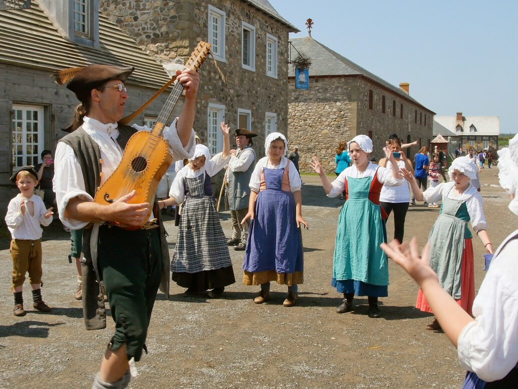 Period actor plays a guitar for others in costume in front of of 18th century building at fortress of louisburg.