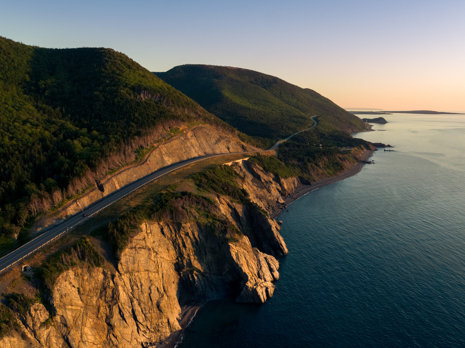 Cap Rouge section of the Cabot Trail with cliffs and ocean lit by the golden glow of a sunset